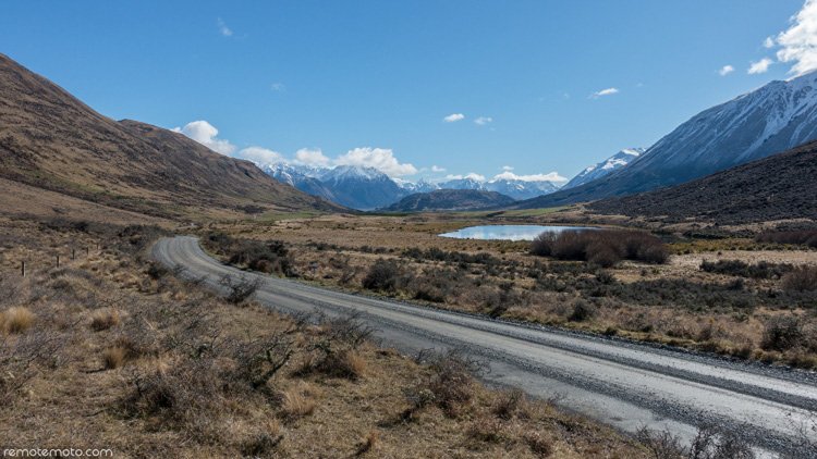 Photo: A gravel road curves right-to-left through a typical yellow-brown NZ high-country valley, past a small lake and towards snow-capped mountains. Blue sky and clouds are reflected in the lake surface.