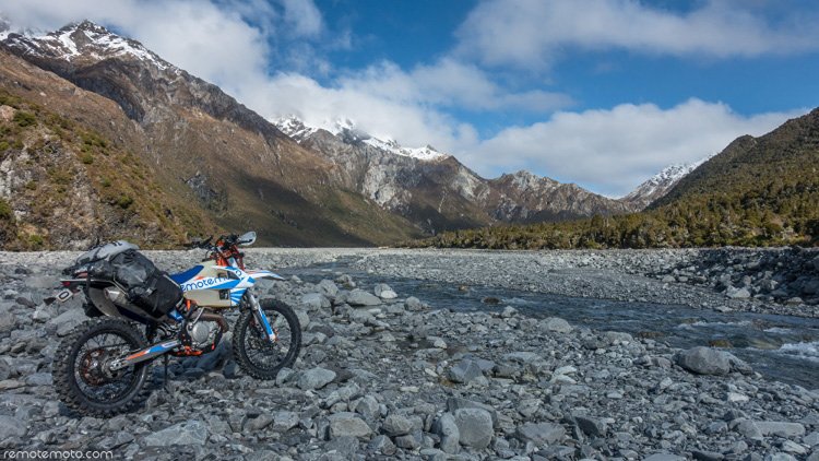 Photo: at the front left a trailbike is parked on a rocky riverbed, with the river flowing from the middle of the photo to the right front corner. The background/upper half of the photo is snow-topped mountains in shades of brown and green, all under a bl