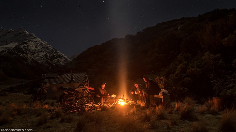 Photo: a night scene. Three people sit around a campfire, bikes and a high-country-hut can be seen behind them. A clear, starry sky is overhead, against which is outlined a bush-covered hill and and a snow-covered mountain can be seen in the distance.